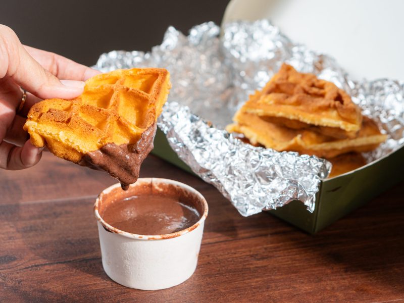 A hand holding a piece of waffle dipped in chocolate sauce, with more waffles in a box lined with foil in the background.